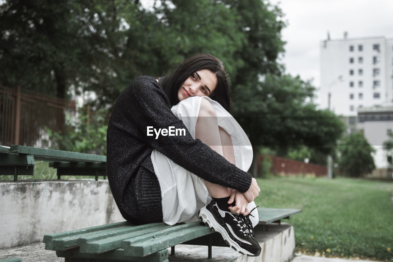 Thoughtful woman hugging knees while sitting on bench at park