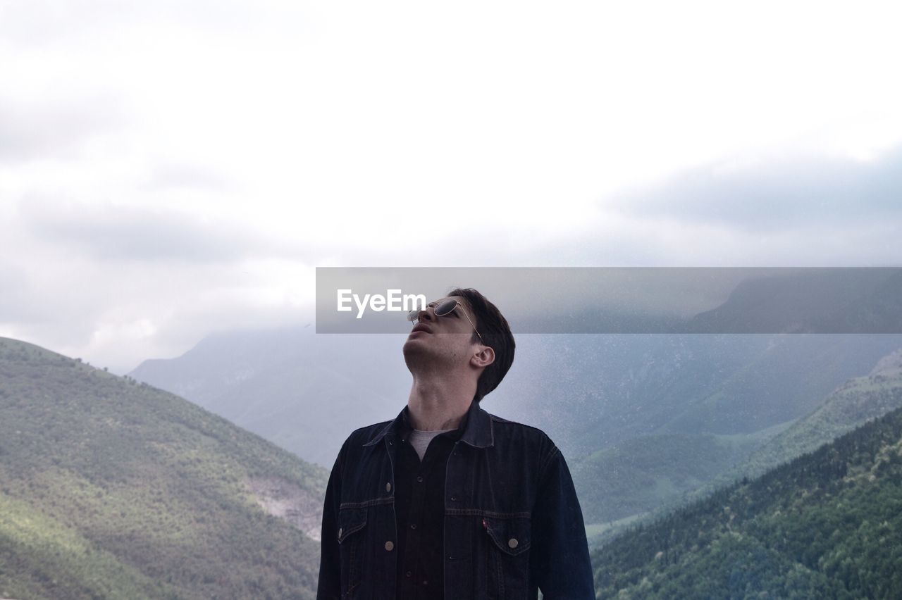 Man in sunglasses standing against mountains during foggy weather