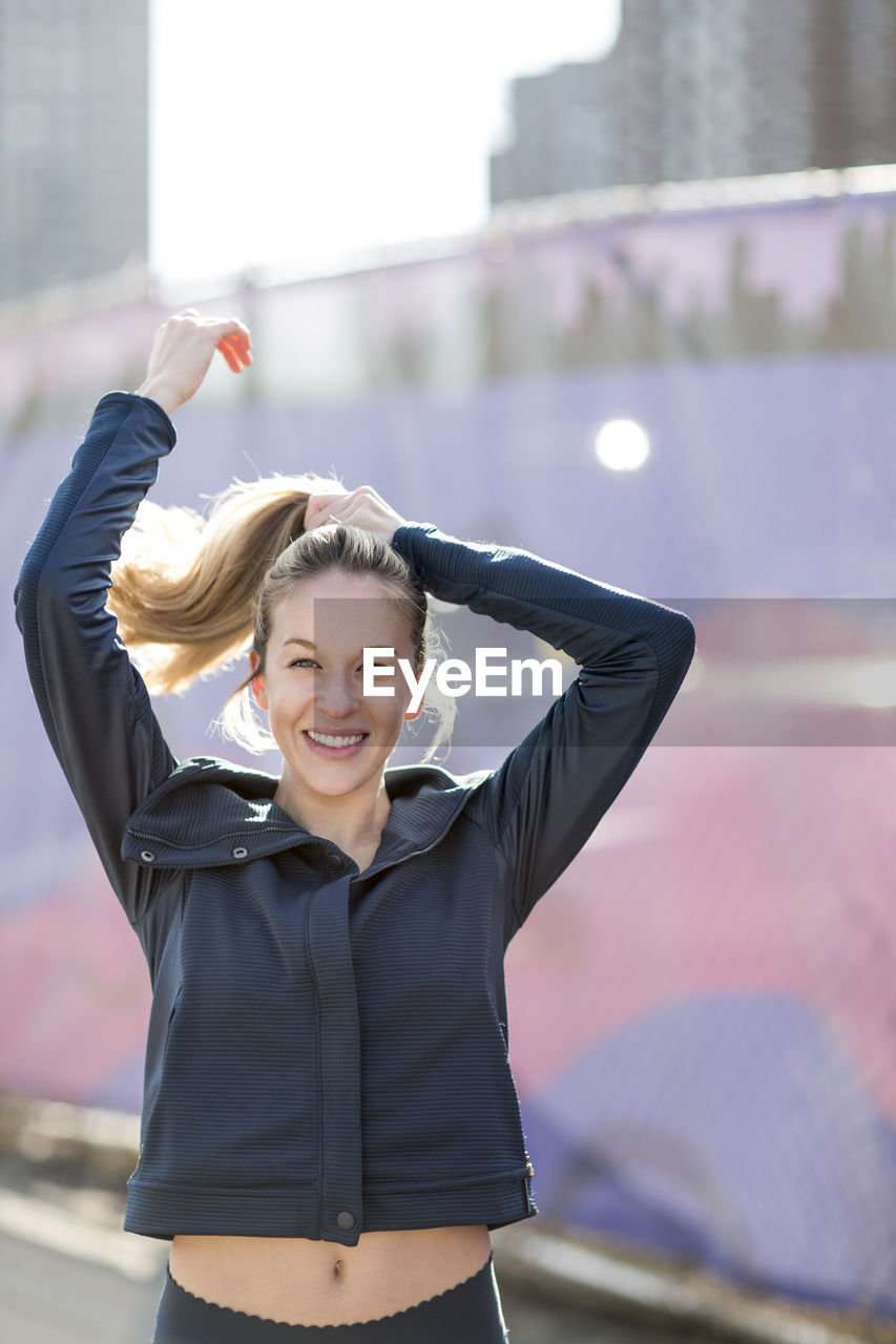 Portrait of confident athlete tying hair while standing by fence during sunny day