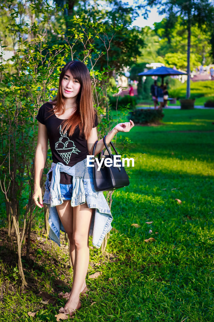 PORTRAIT OF SMILING YOUNG WOMAN STANDING AGAINST GREEN PLANTS