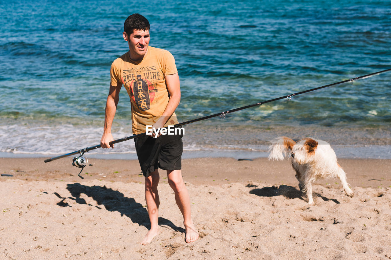 Man holding fishing rod while standing with dog at beach