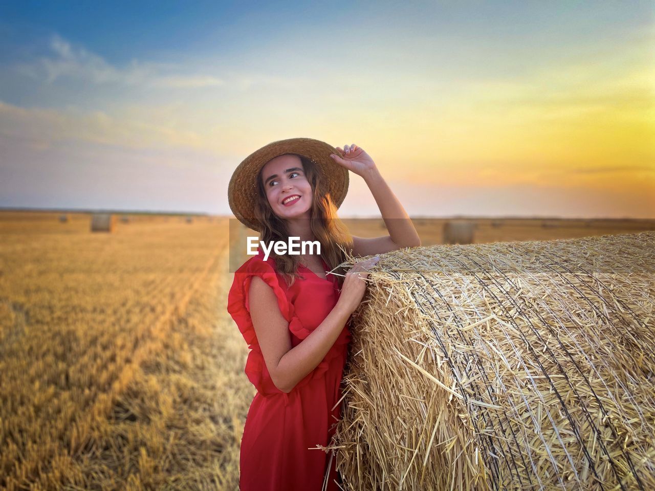 Portrait of young woman wearing straw hat and red dress standing near a hay bale at sunset