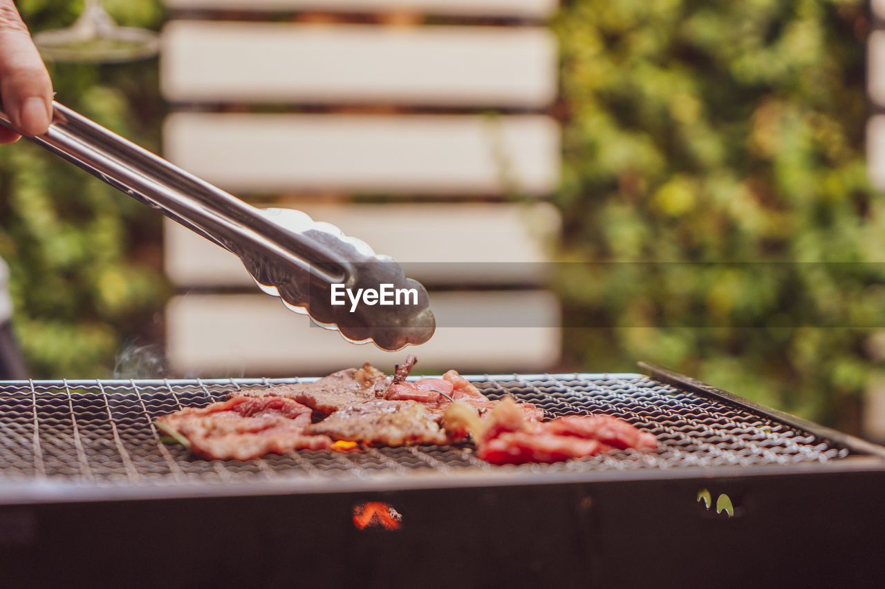 Cropped hand using meat tongs to grill slices of premium meat on the stovetop at 
 outdoor party. 