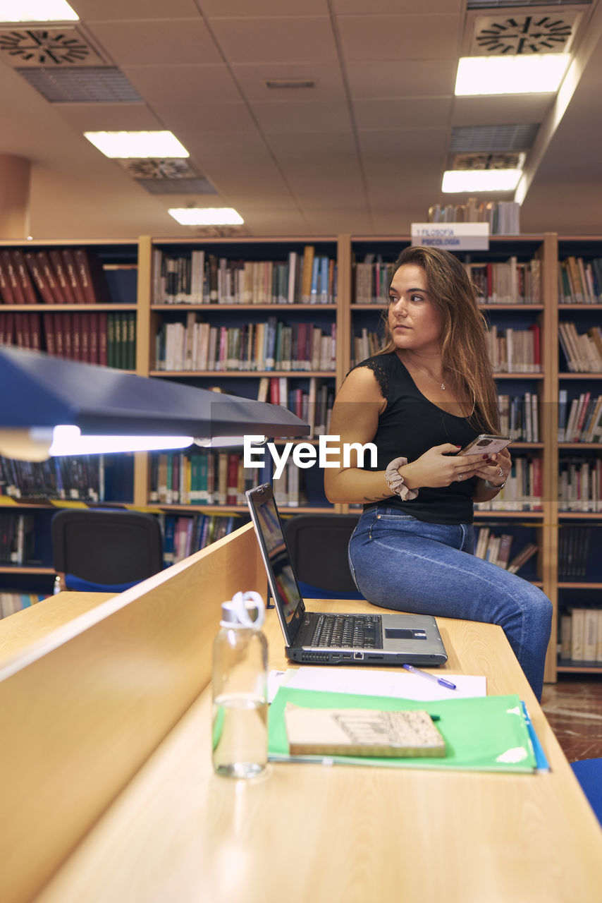 A young girl with a mobile phone is sitting at a library table