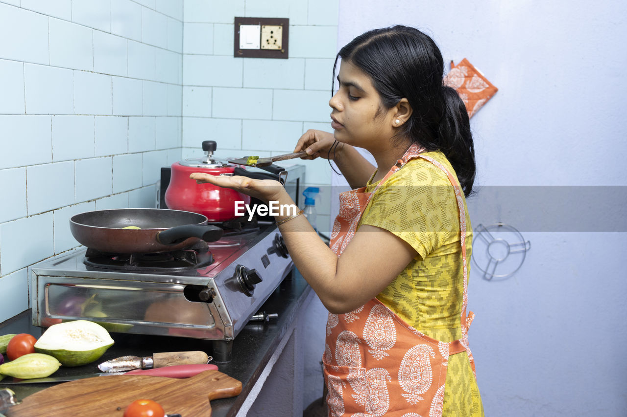 A pretty indian young woman wearing apron cooking and tasting food in domestic kitchen on gas stove