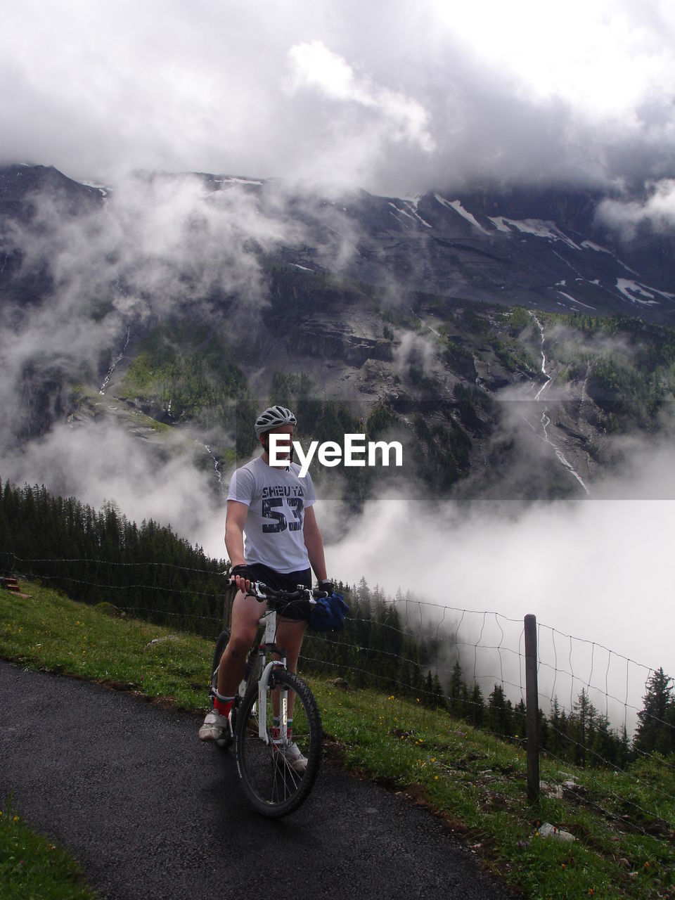 Full length of young man with mountain bike standing against cloudy sky