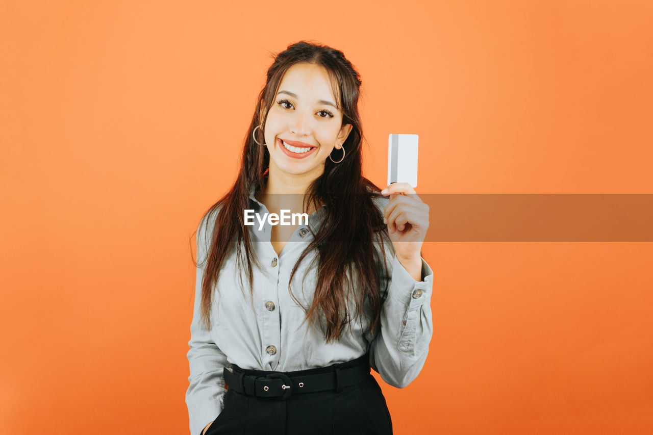 one person, adult, smiling, women, colored background, portrait, studio shot, happiness, young adult, orange color, technology, long hair, wireless technology, indoors, looking at camera, copy space, smartphone, photo shoot, portable information device, hairstyle, communication, holding, front view, emotion, mobile phone, person, standing, clothing, cheerful, waist up, orange background, brown hair, business, fashion, lifestyles, female, telephone, positive emotion, human face, casual clothing, enjoyment, smile