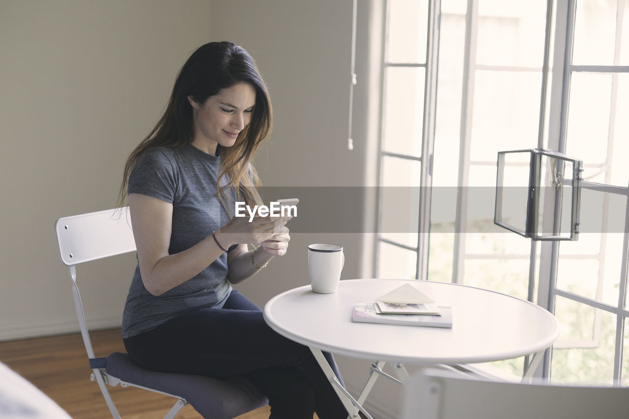 Woman text messaging on smart phone while having coffee at table by window in living room