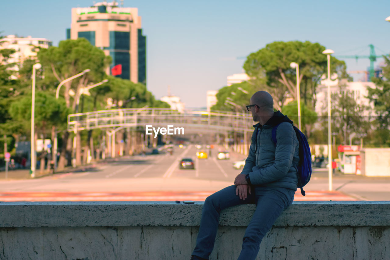 MAN SITTING ON RETAINING WALL AGAINST CITY IN BACKGROUND