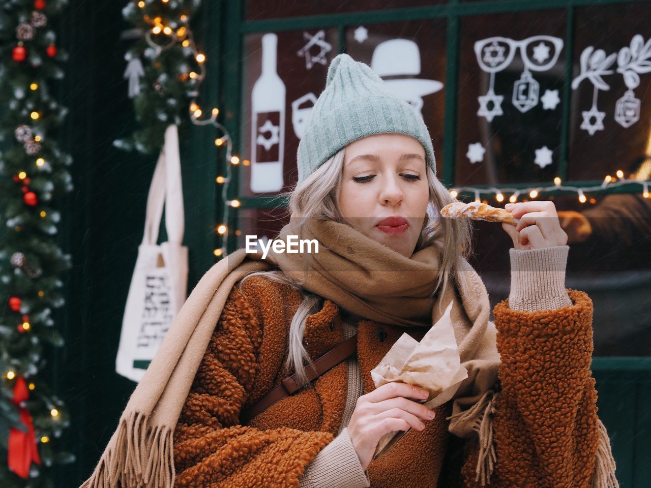 Young woman with blonde hair wearing brown fur coat eating sweet food on the christmas market. 