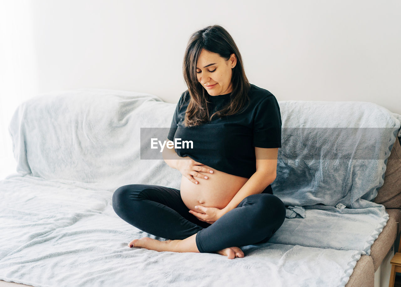 A middle-aged pregnant woman carefully hugs her belly while sitting on the couch at home.