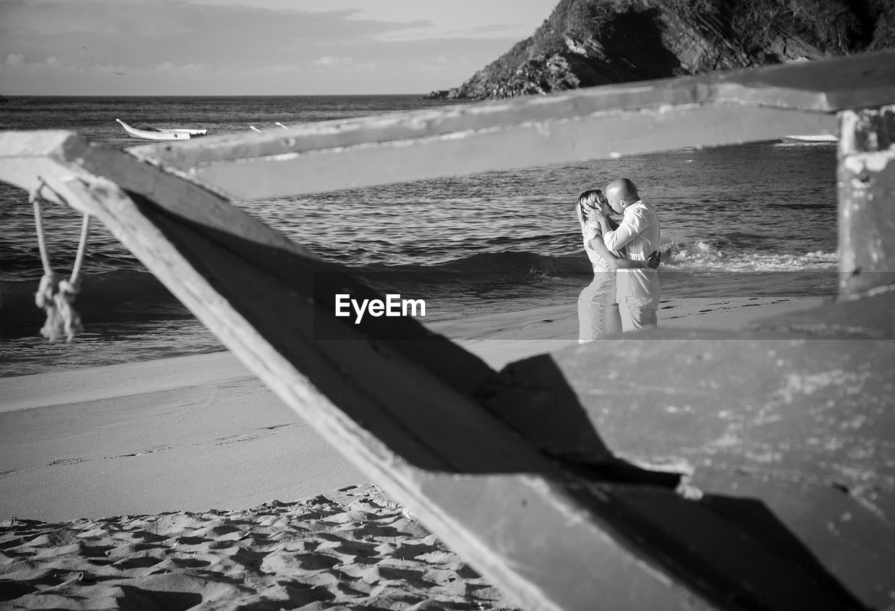 A loving couple kiss on a beach, close-up view of a boat that frames them