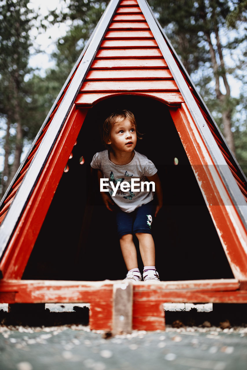 Child playing in a red woden tent at the natural kids playground surrounded with pine tree forest.