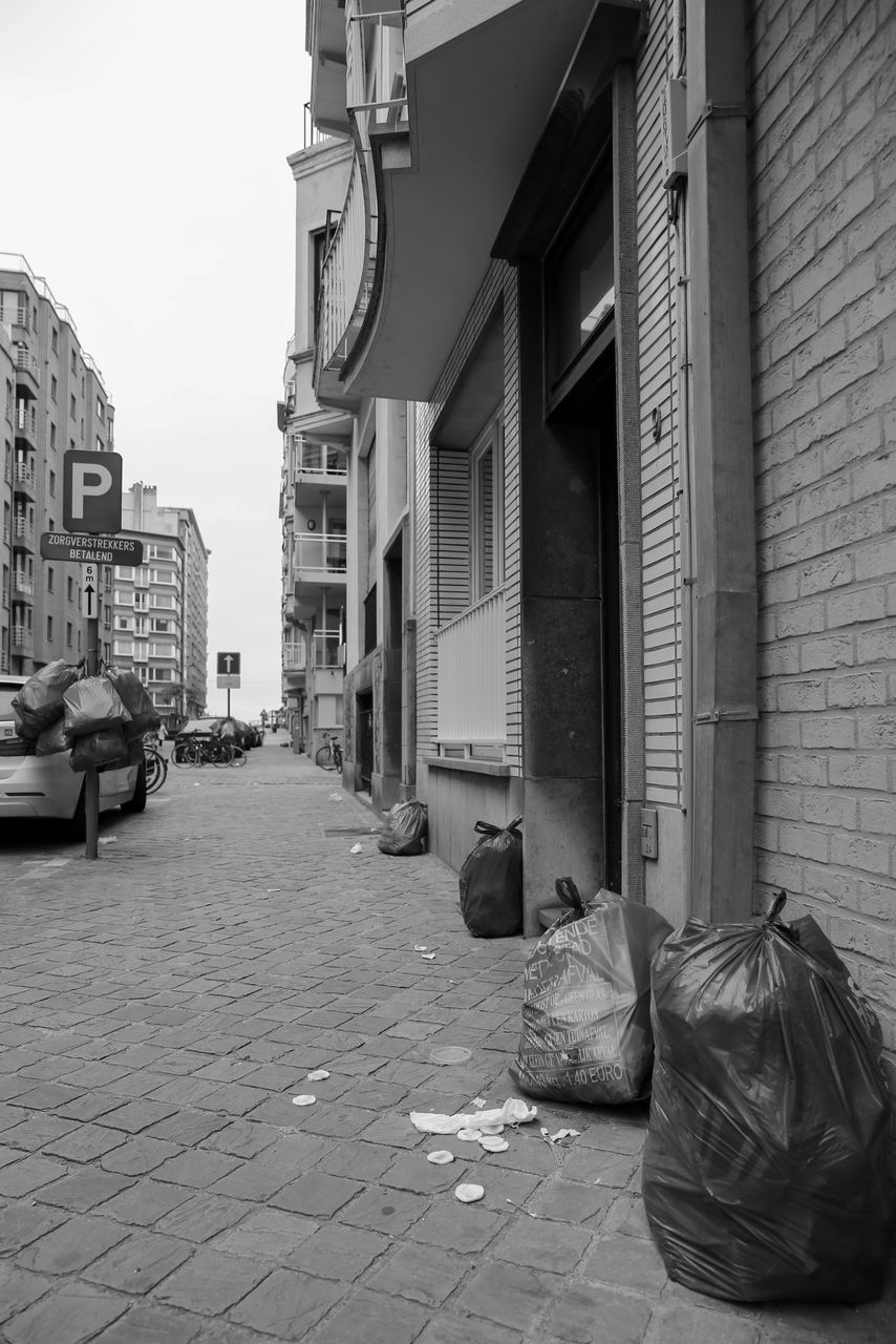 road, street, architecture, building exterior, city, built structure, infrastructure, alley, black and white, black, urban area, monochrome, building, monochrome photography, lane, footpath, day, white, transportation, cobblestone, mode of transportation, sidewalk, residential district, no people, road surface, car, waste container, outdoors, city life, city street, homelessness, motor vehicle