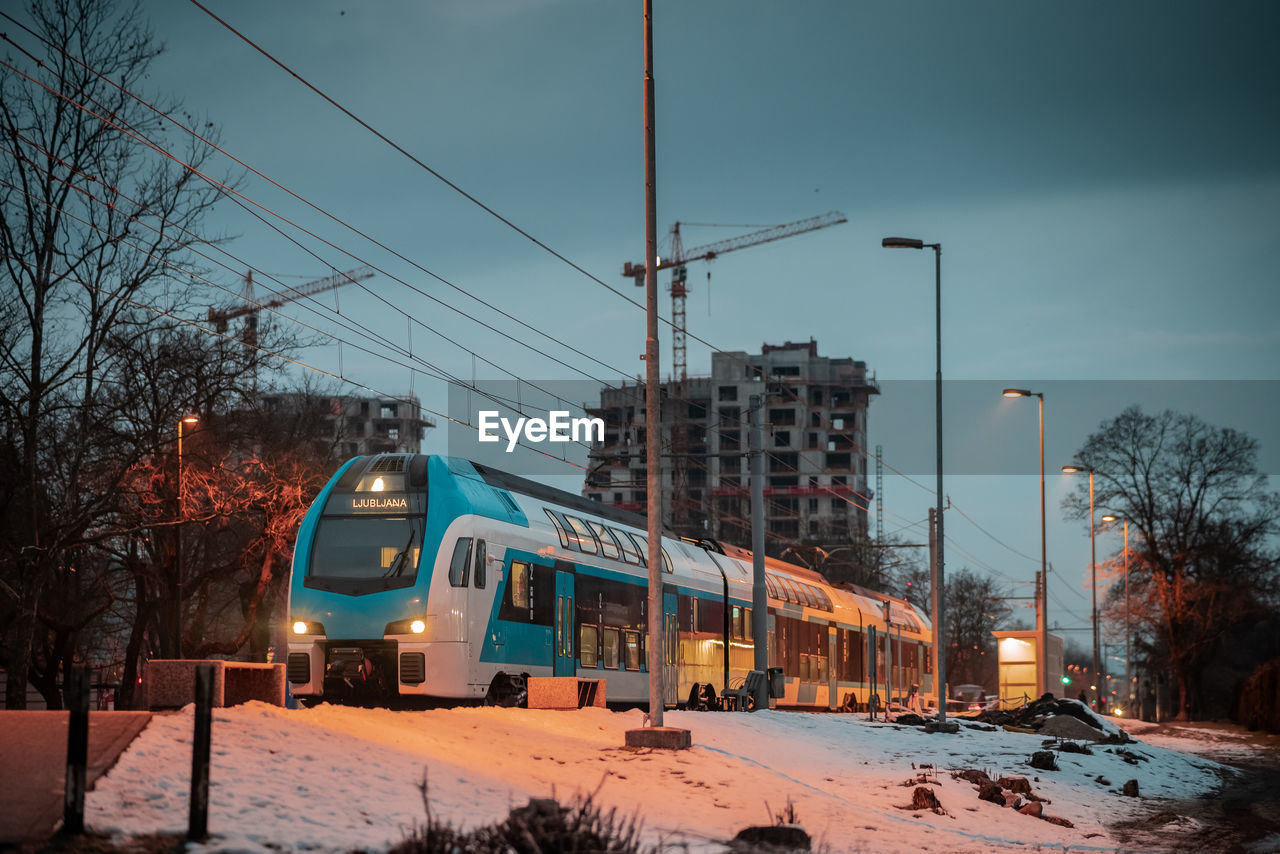snow, winter, transportation, mode of transportation, cold temperature, architecture, sky, night, nature, evening, electricity, urban area, city, tree, building exterior, street, built structure, train, vehicle, rail transportation, no people, illuminated, land vehicle, street light, road, motor vehicle, bare tree, cityscape, outdoors, cable, plant, railroad track, dusk, car, public transportation, lighting, travel, transport, track, residential area, electrical supply