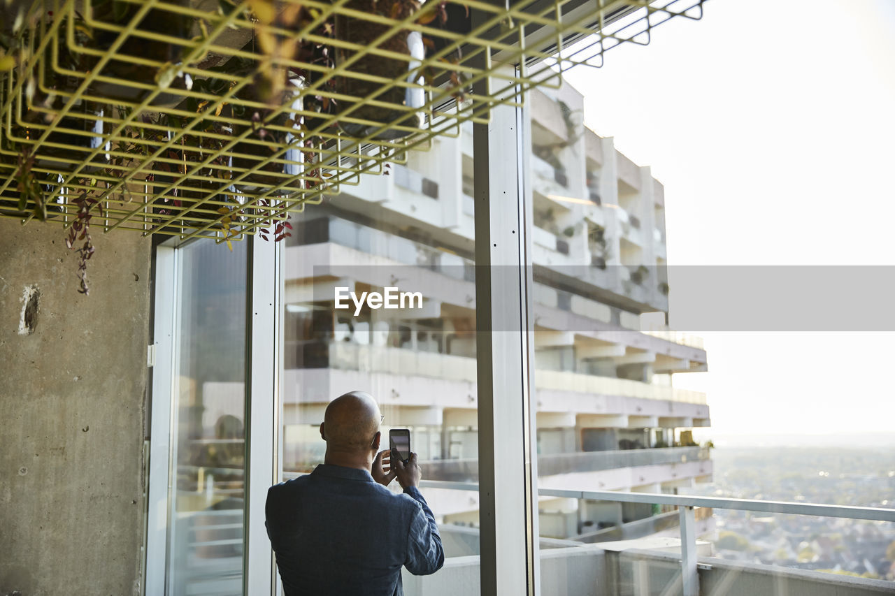 Mature man looking out of window while taking photo on smart phone