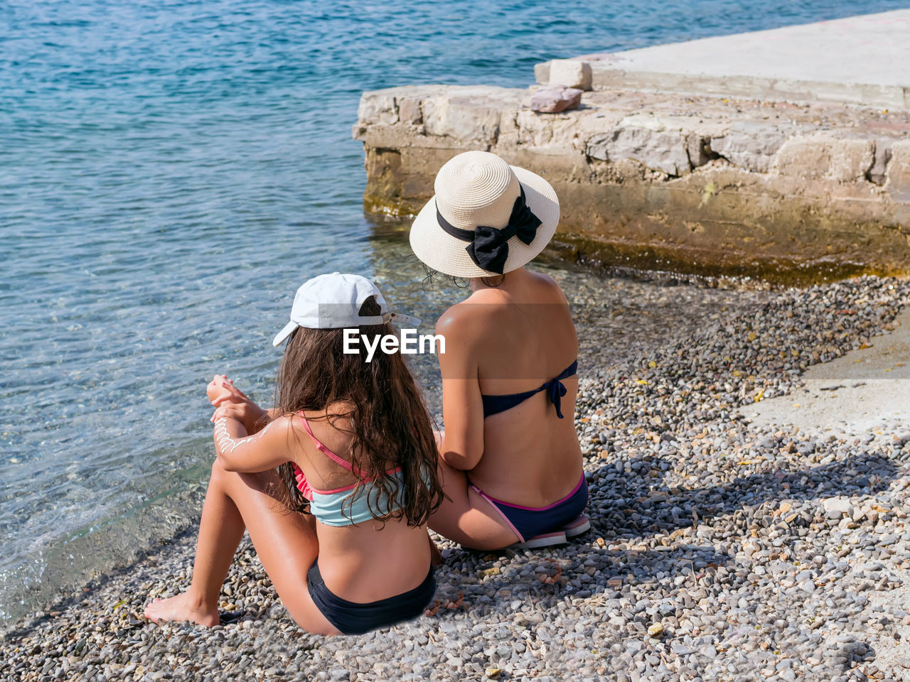 Unrecognizable caucasian mom and her daughter wearing swimsuit are sitting on the beach.