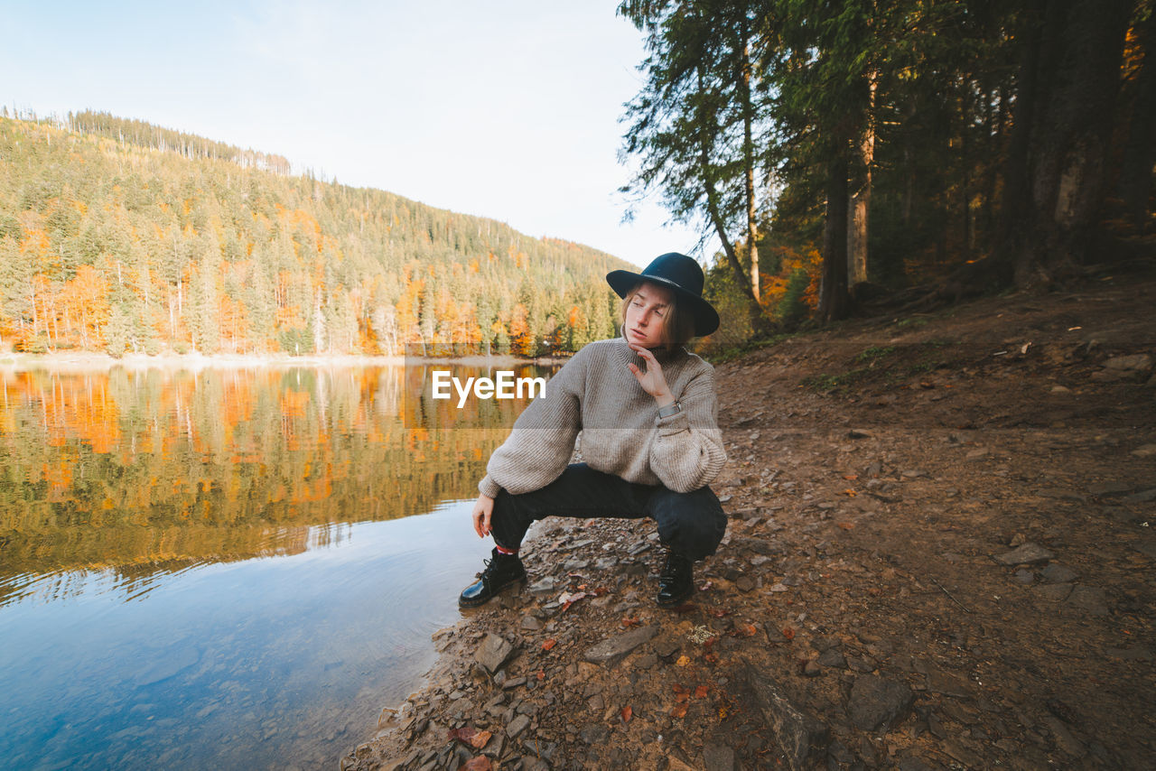 Young cool female tourist in trendy apparel looking away from dry coast against transparent water reflecting autumn trees