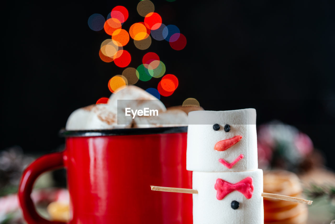 Red enamel mug of hot chocolate with marshmallow snowman on christmas background