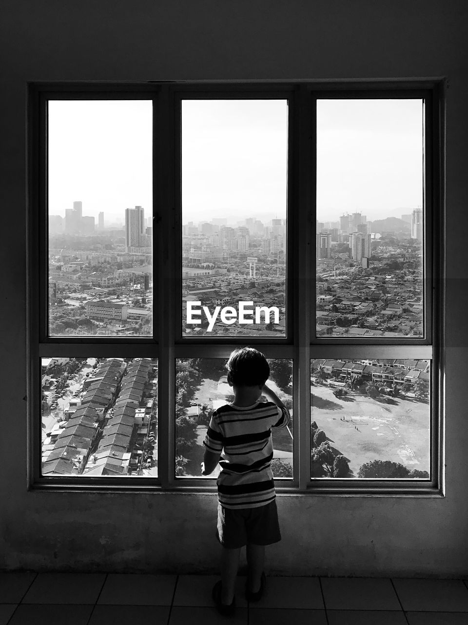 Rear view of a small boy standing by window looking out at the city