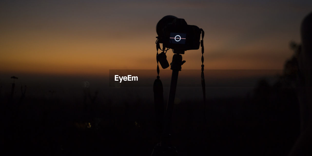 SILHOUETTE OF CAMERA ON FIELD AGAINST ORANGE SKY