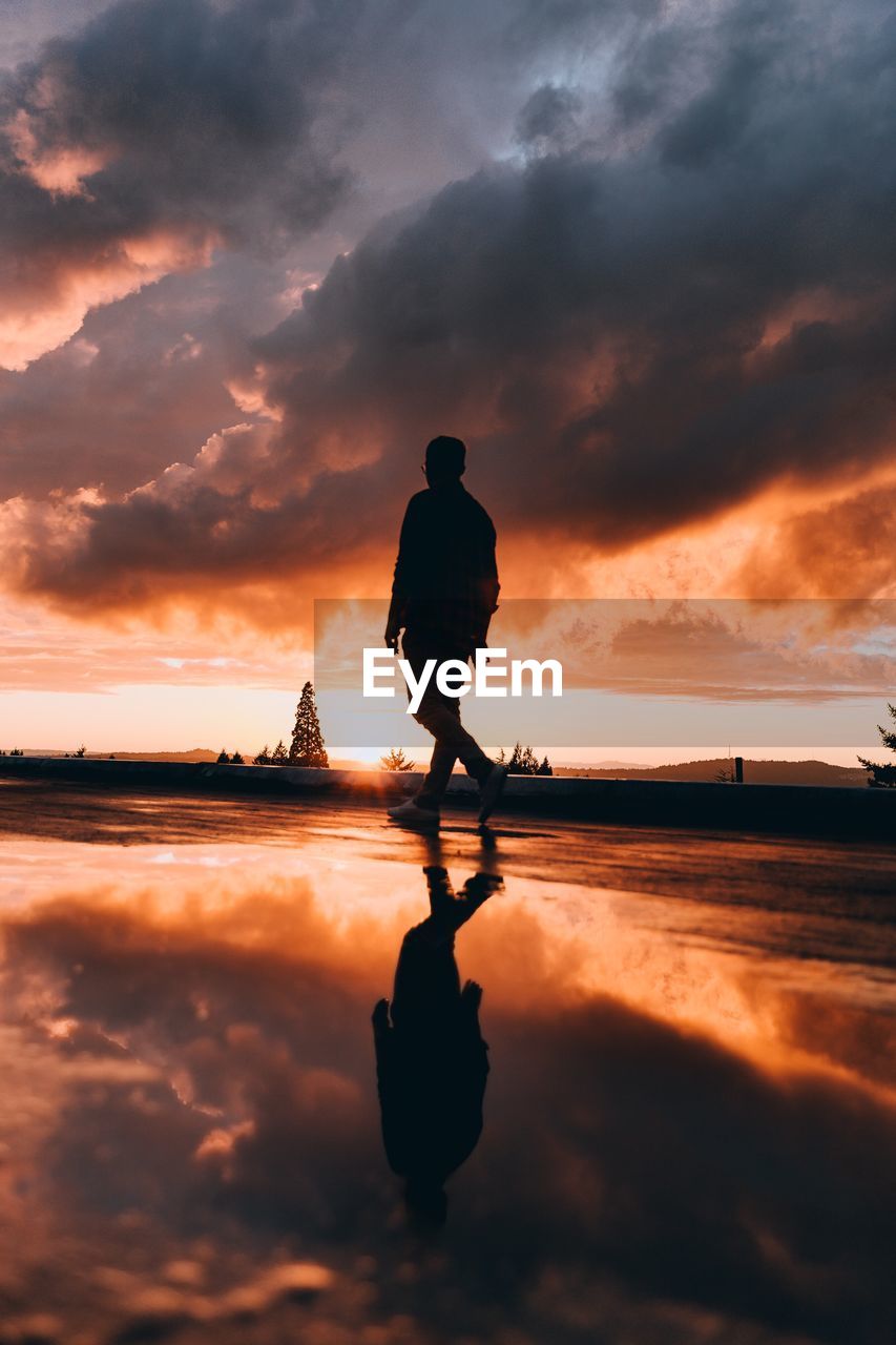 Reflection of silhouette man walking on puddle against cloudy sky