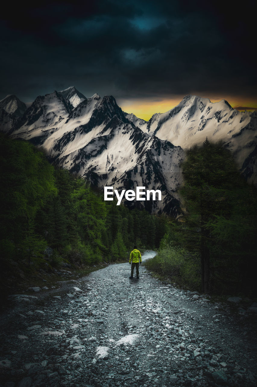 Rear view of man walking on dirt footpath leading towards snowcapped mountains at night