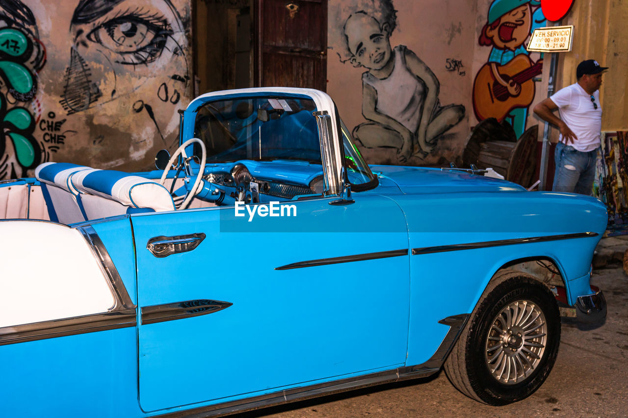 car, mode of transportation, vehicle, transportation, land vehicle, motor vehicle, antique car, vintage car, automobile, men, city, architecture, adult, travel, retro styled, day, two people, street, person, driving, blue, outdoors, graffiti