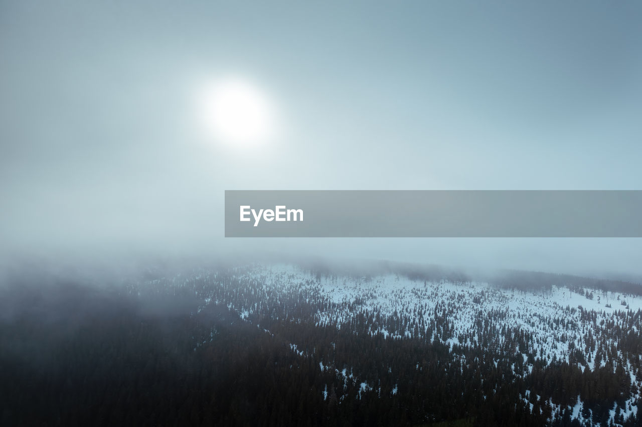 Aerial view of snowy mountain range in cloud with sun shining through during winter day.