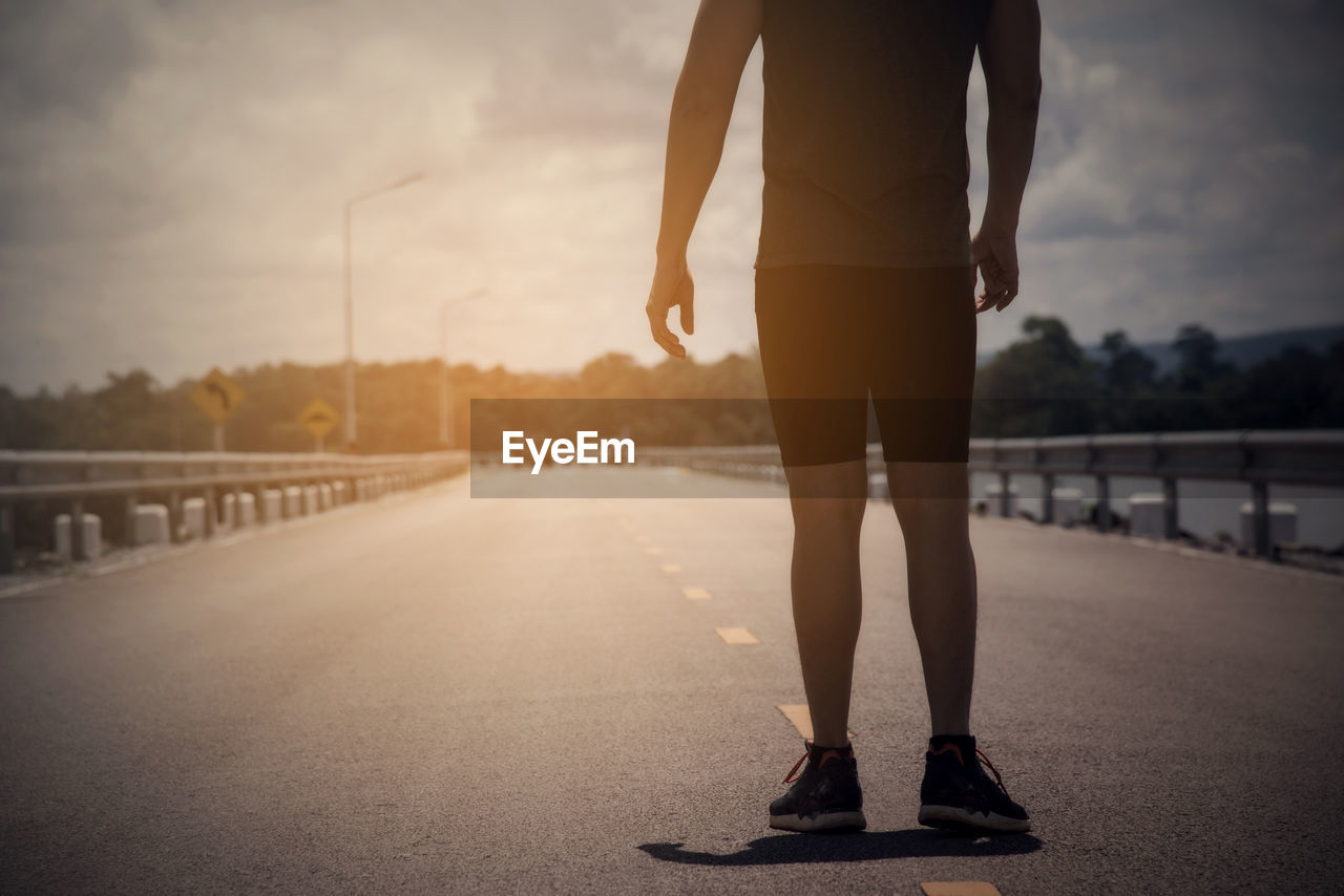 LOW SECTION OF WOMAN STANDING ON ROAD AGAINST SKY DURING SUNSET