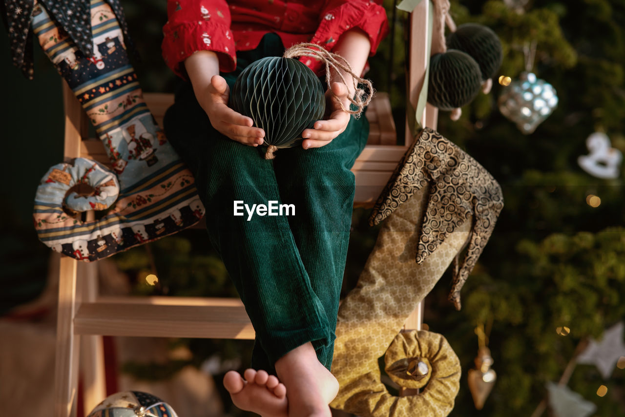 Barefoot boy in an elf costume holds craft paper deep green christmas tree ball
