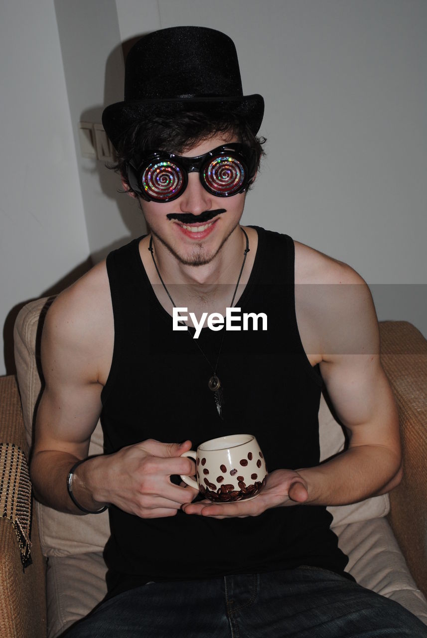 Man with coffee cup wearing funny goggles while sitting on sofa at home