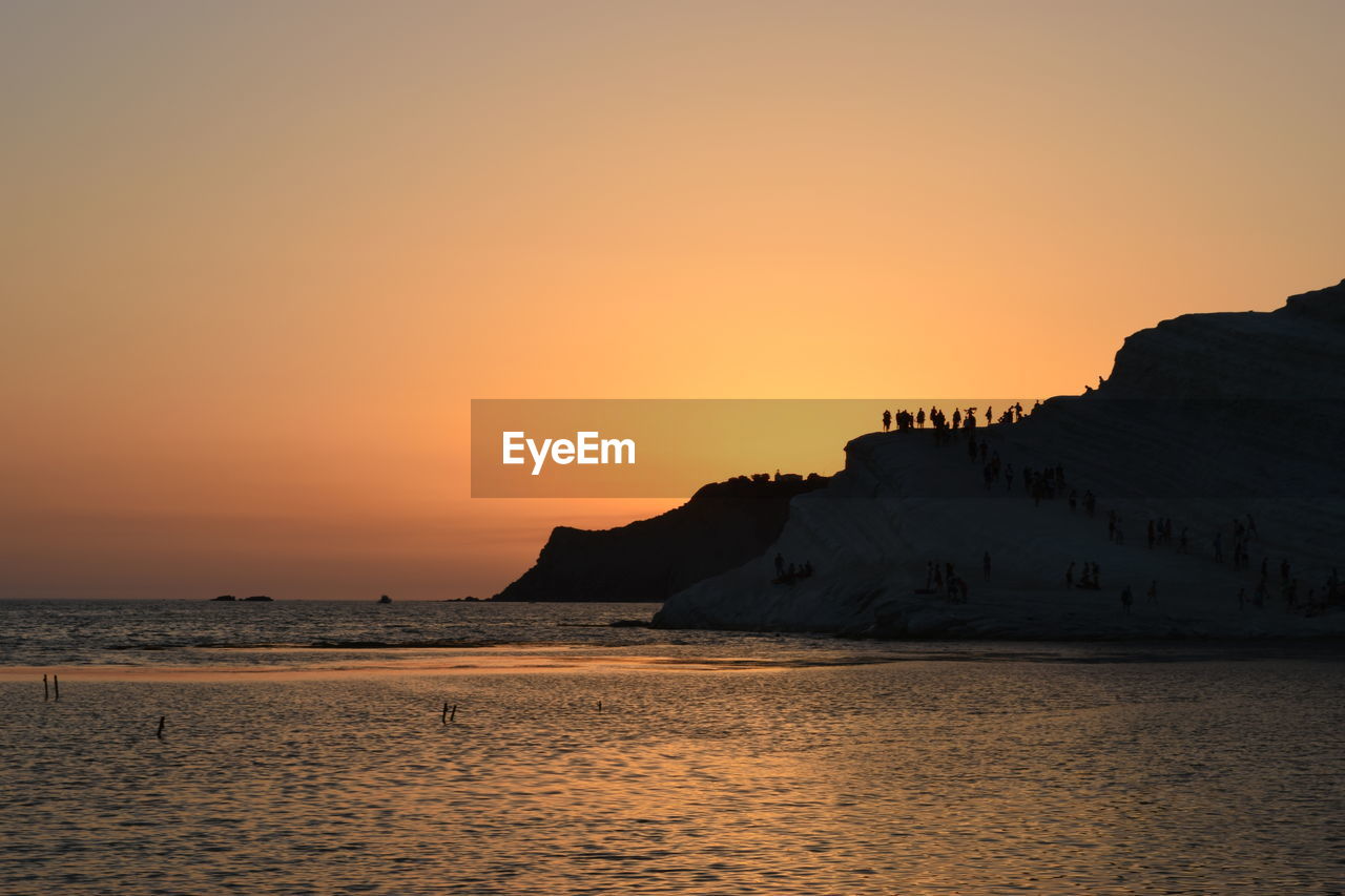 SILHOUETTE MOUNTAIN BY SEA AGAINST ORANGE SKY