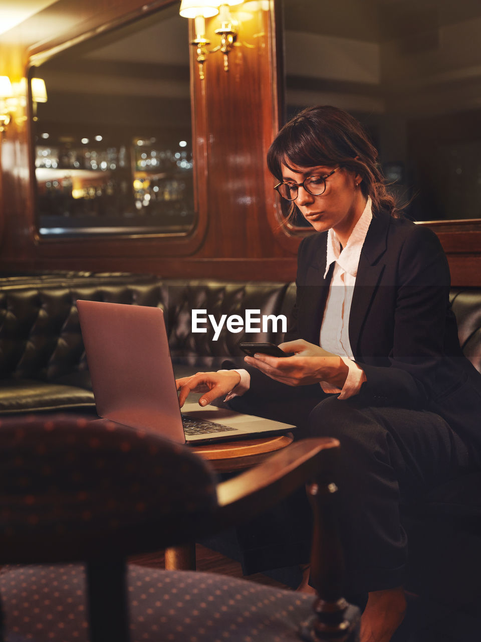 Smart business woman in glasses typing on laptop and checking information in mobile phone comfortably sitting in black leather sofa in cafe