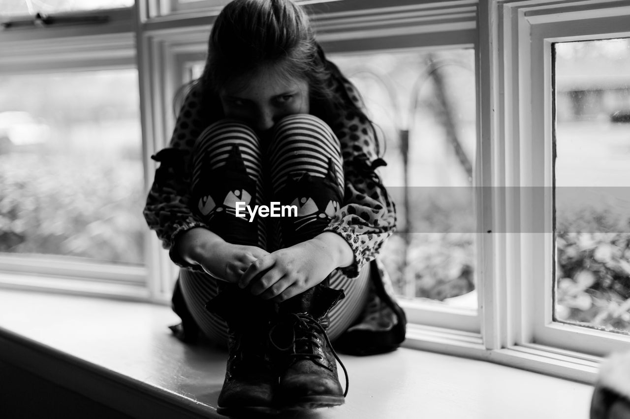 Close-up of girl hugging knees while sitting on window sill