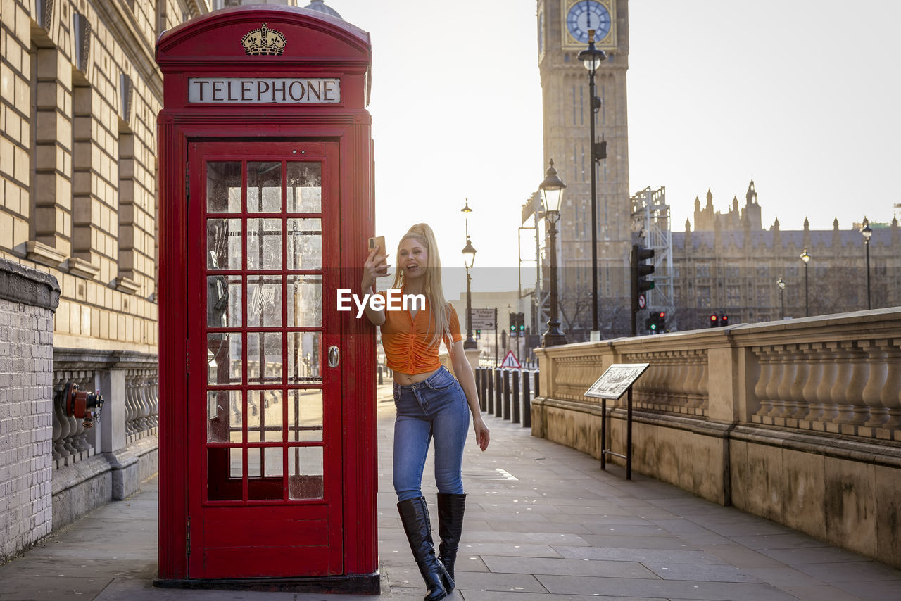 Young woman taking selfie by telephone booth on sunny day