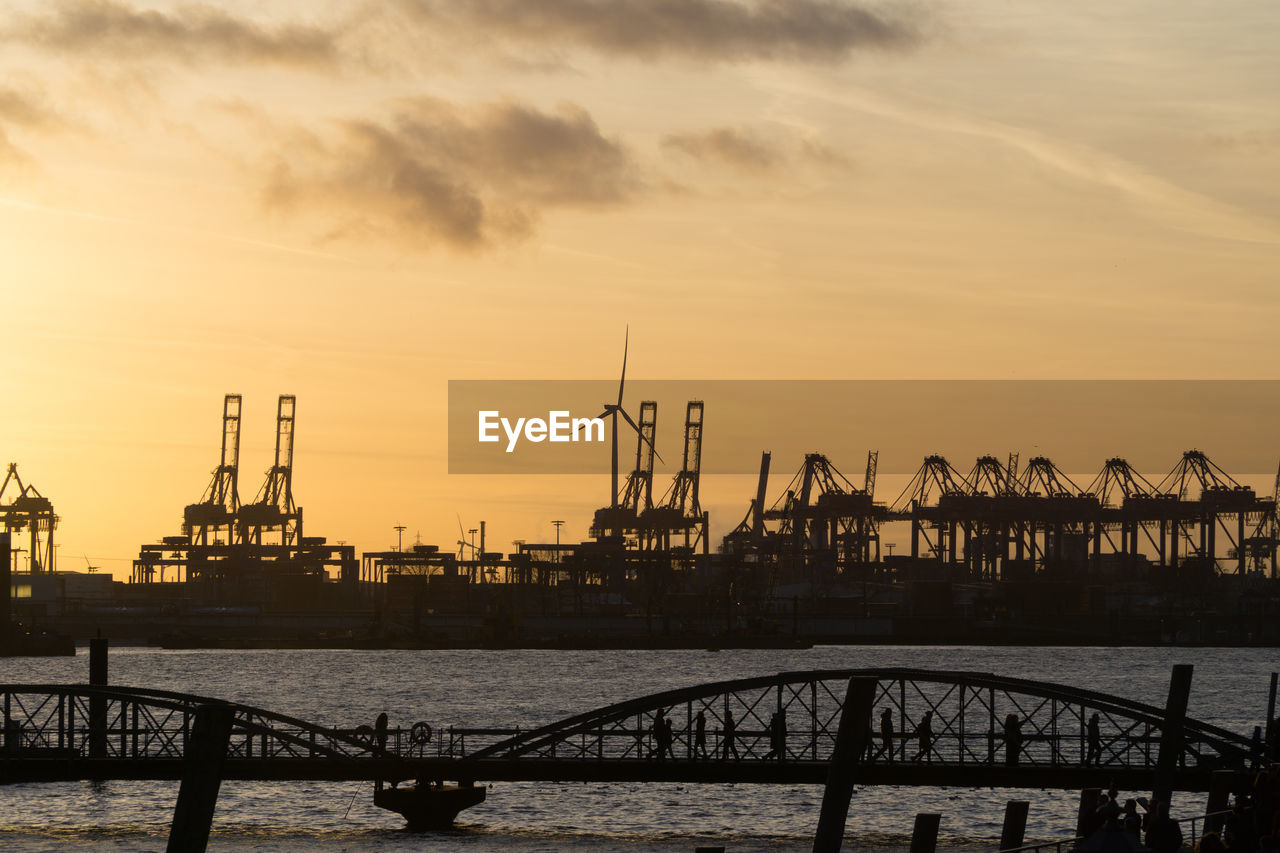 Silhouette cranes at harbor against sky during sunset