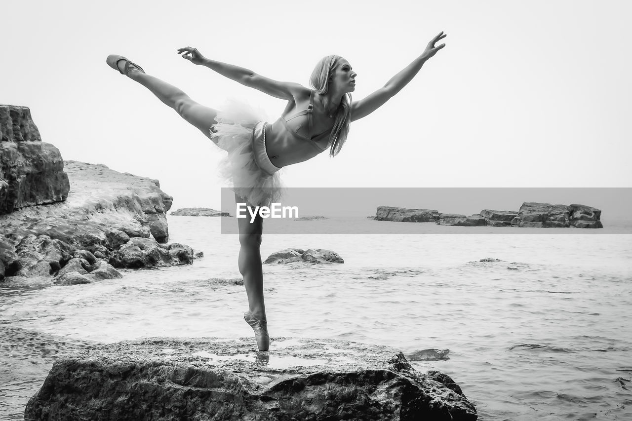 Full length of young woman dancing rock by sea against clear sky