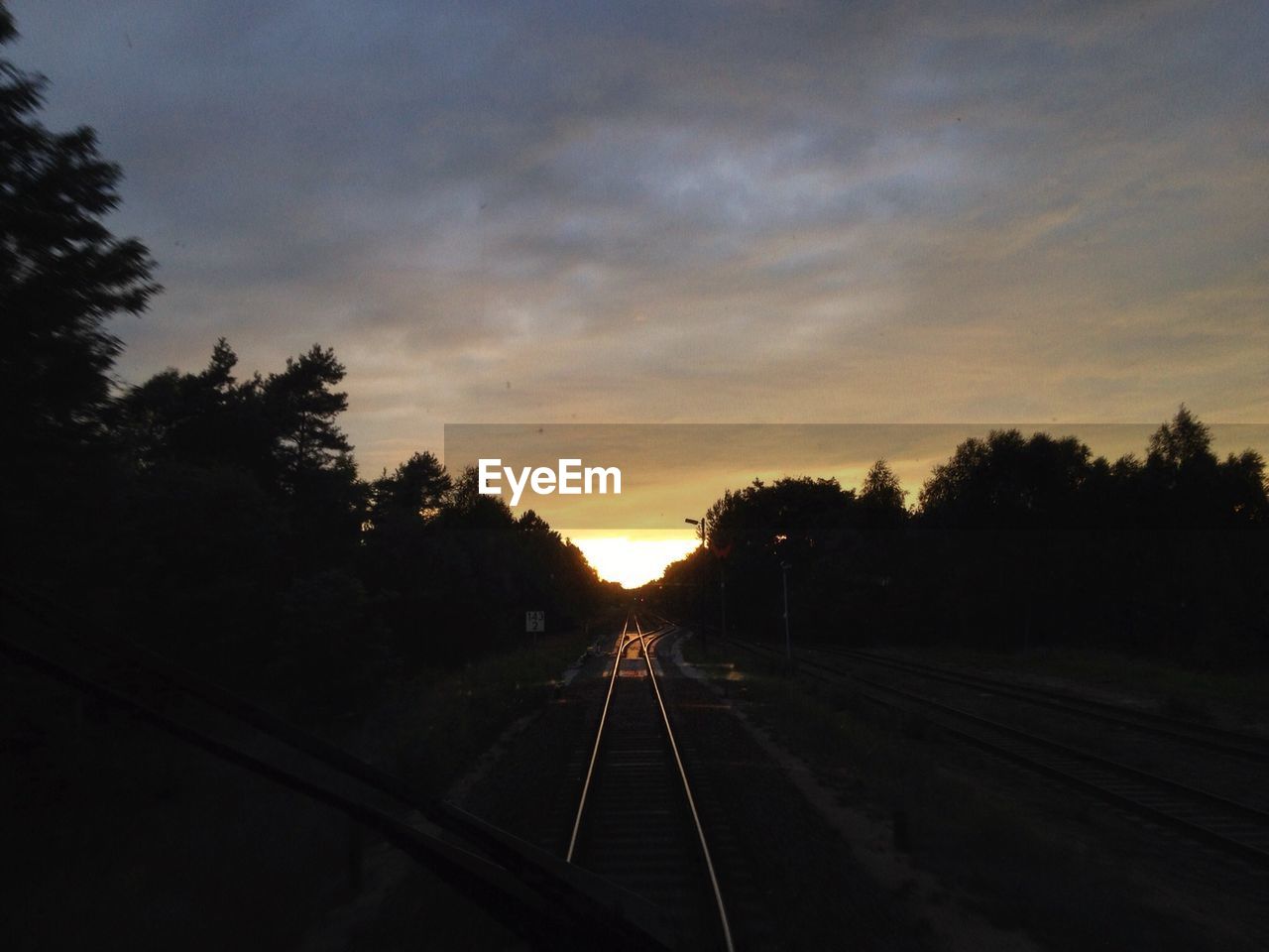 Empty railroad track amidst silhouette trees against cloudy sky during sunset