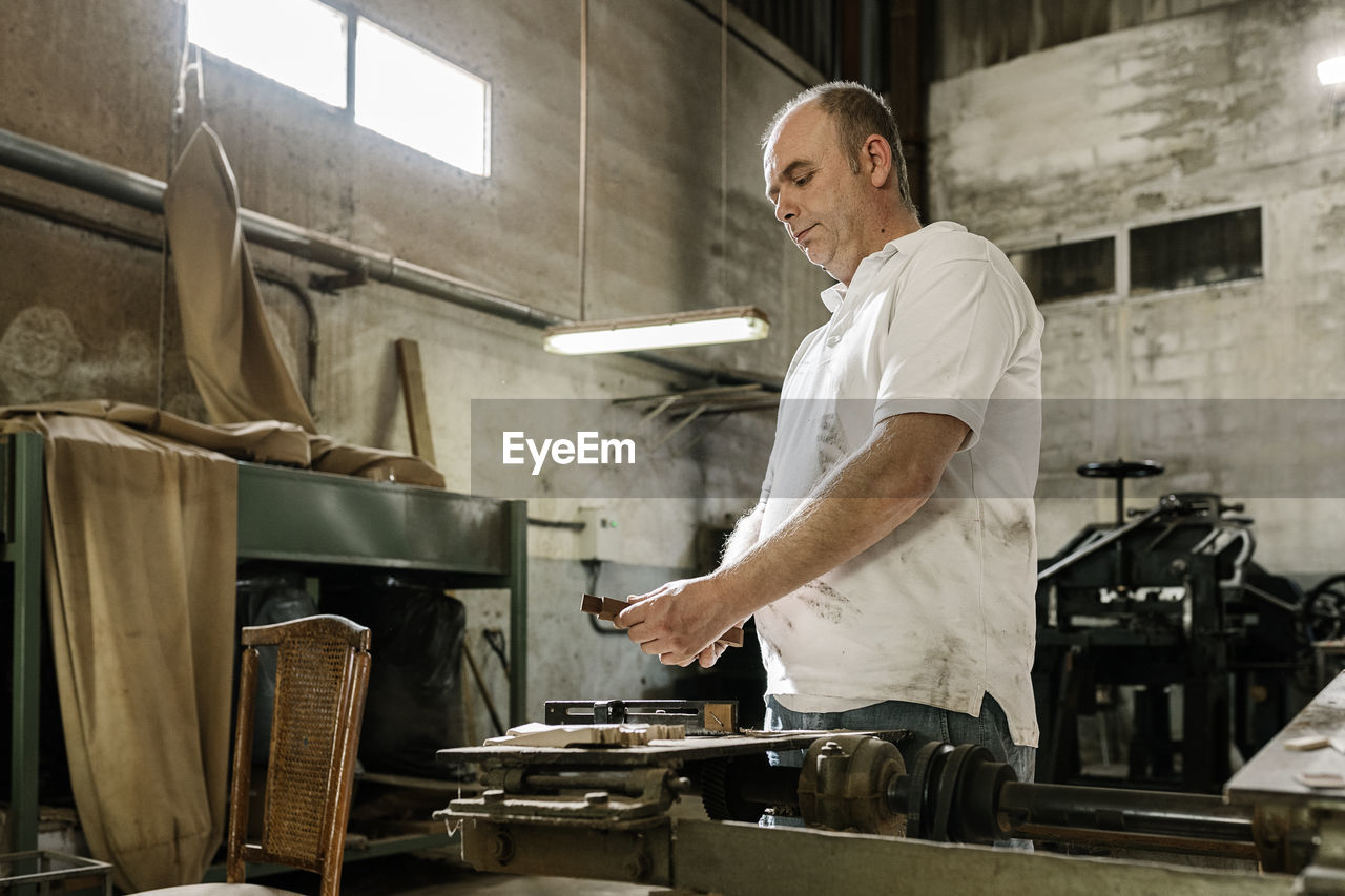 Low angle side view of concentrated male woodworker creating wooden details while standing in grungy garage at daytime