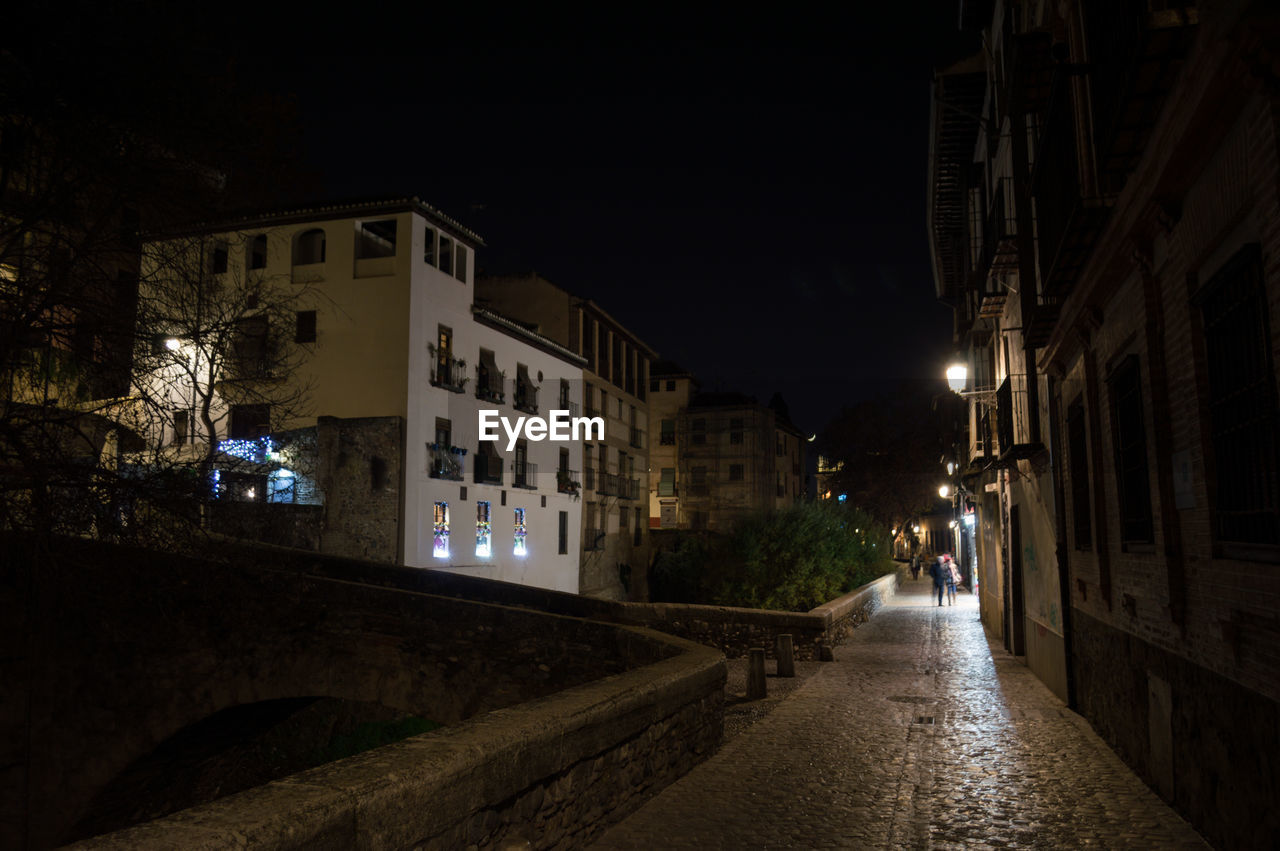 STREET AMIDST BUILDINGS AGAINST SKY AT NIGHT