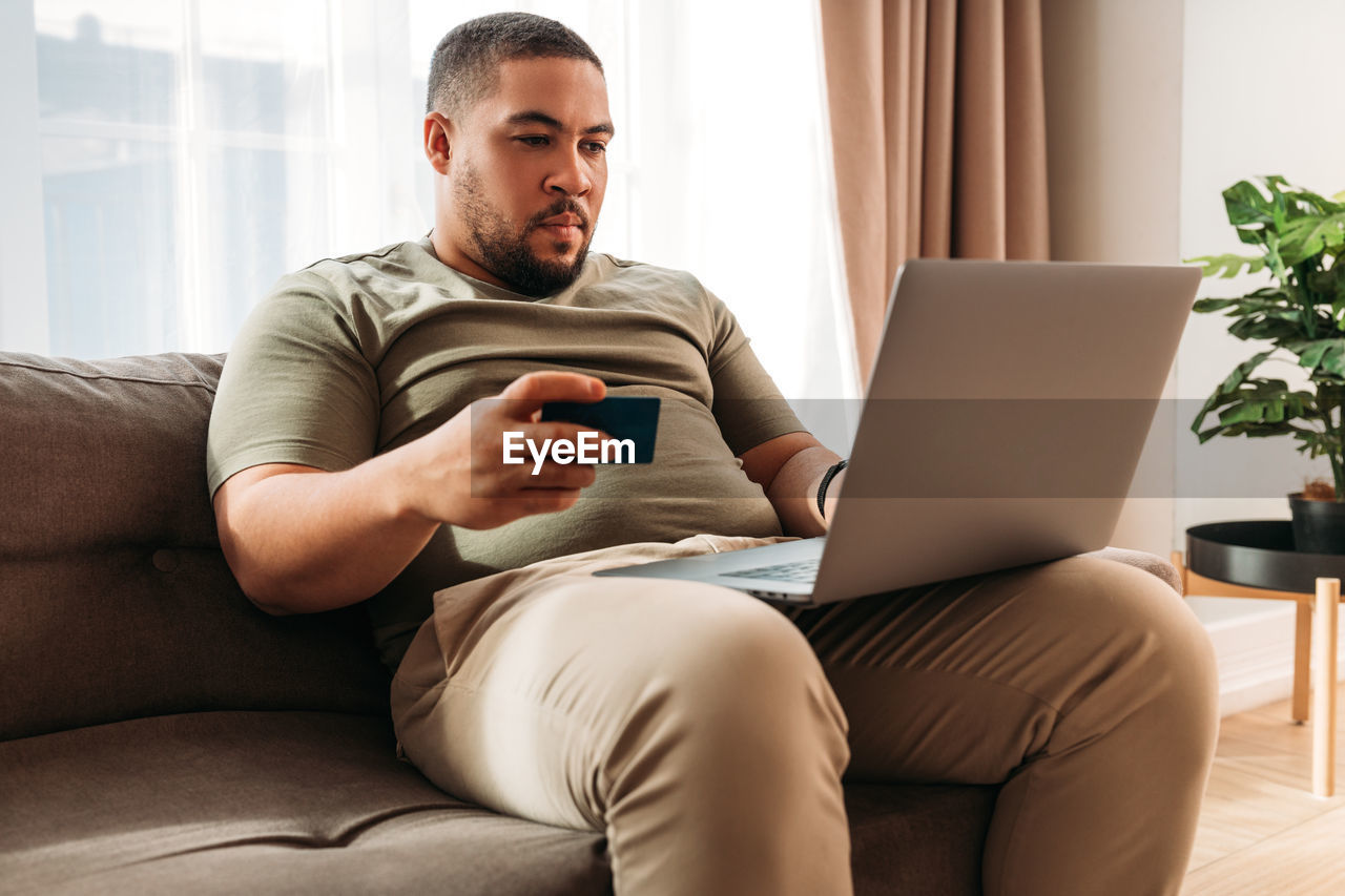 Smiling man using laptop while holding credit card sitting on sofa at home