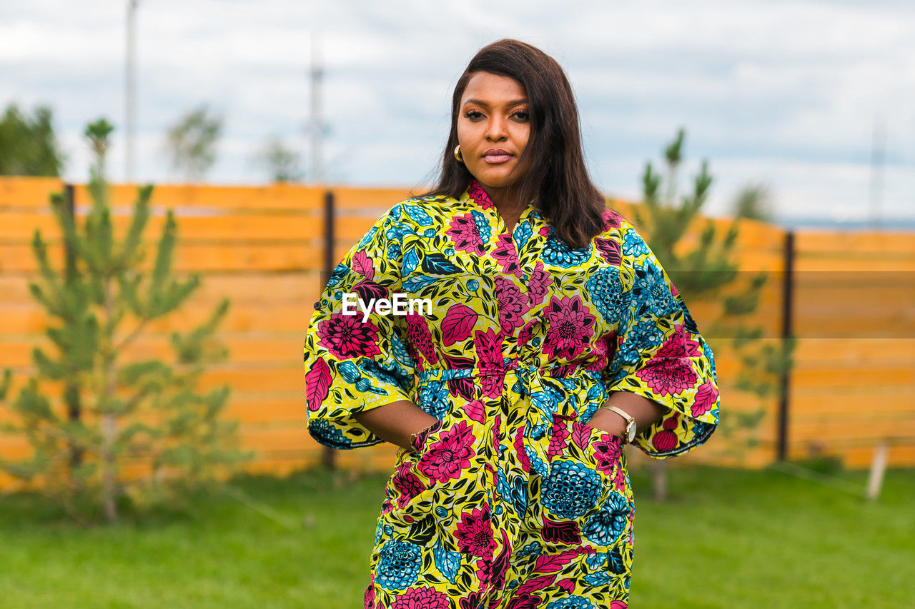 one person, women, spring, adult, yellow, smiling, standing, portrait, dress, young adult, nature, happiness, flower, clothing, hairstyle, looking at camera, lifestyles, multi colored, emotion, grass, front view, fashion, pattern, plant, female, floral pattern, focus on foreground, outdoors, sky, brown hair, photo shoot, three quarter length, cheerful, architecture, long hair, leisure activity, day, waist up, casual clothing, green, person, sleeve, copy space