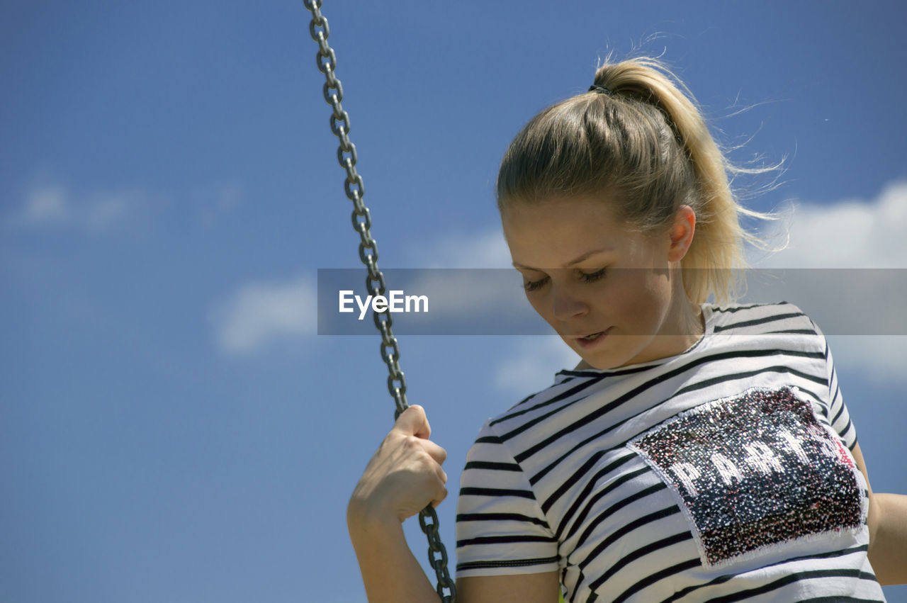 Low angle view of young woman looking down while swinging against sky