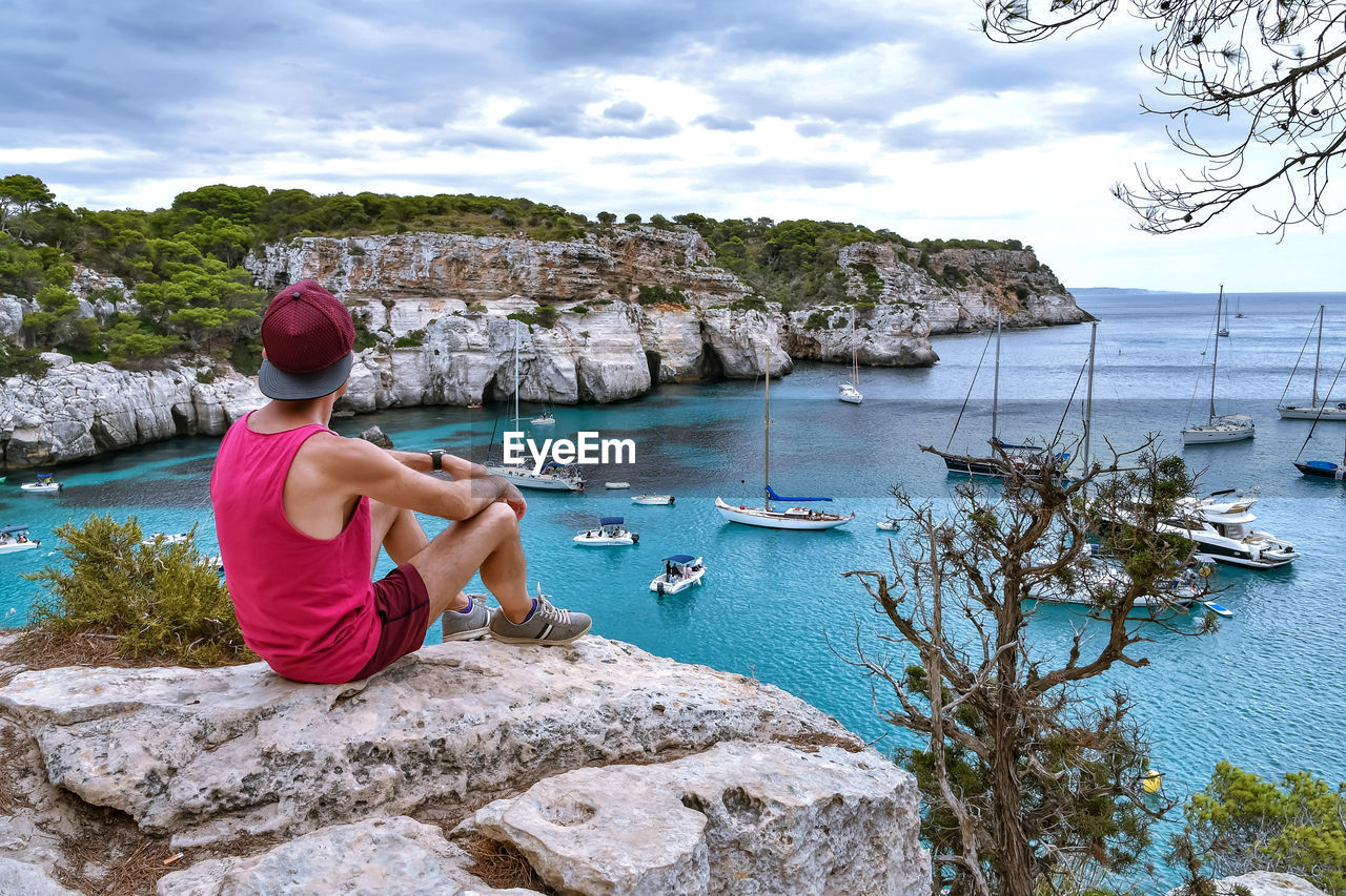 Rear view of man sitting on rock looking at sea against sky