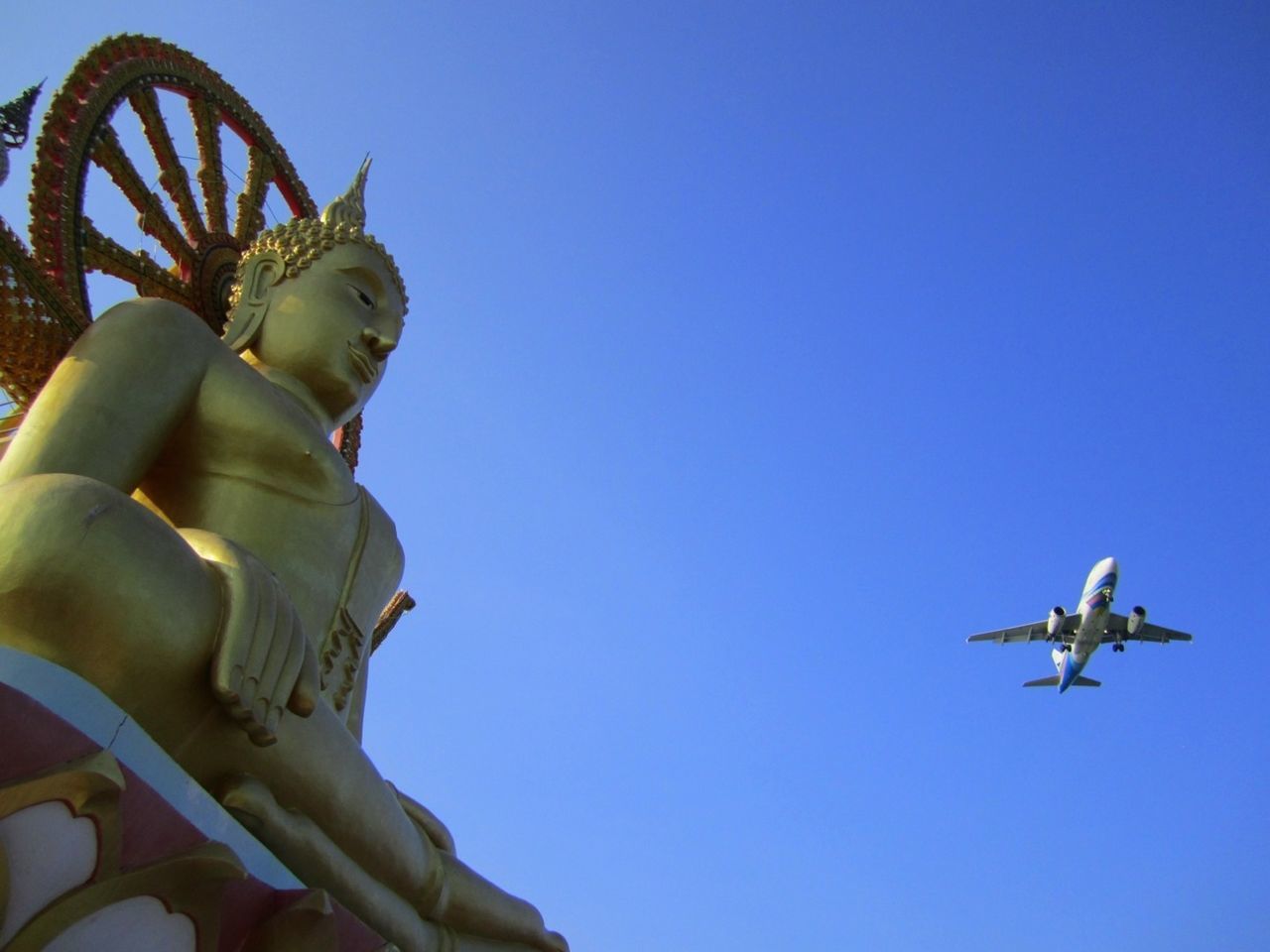 Low angle view of buddha statue against airplane in clear blue sky on sunny day