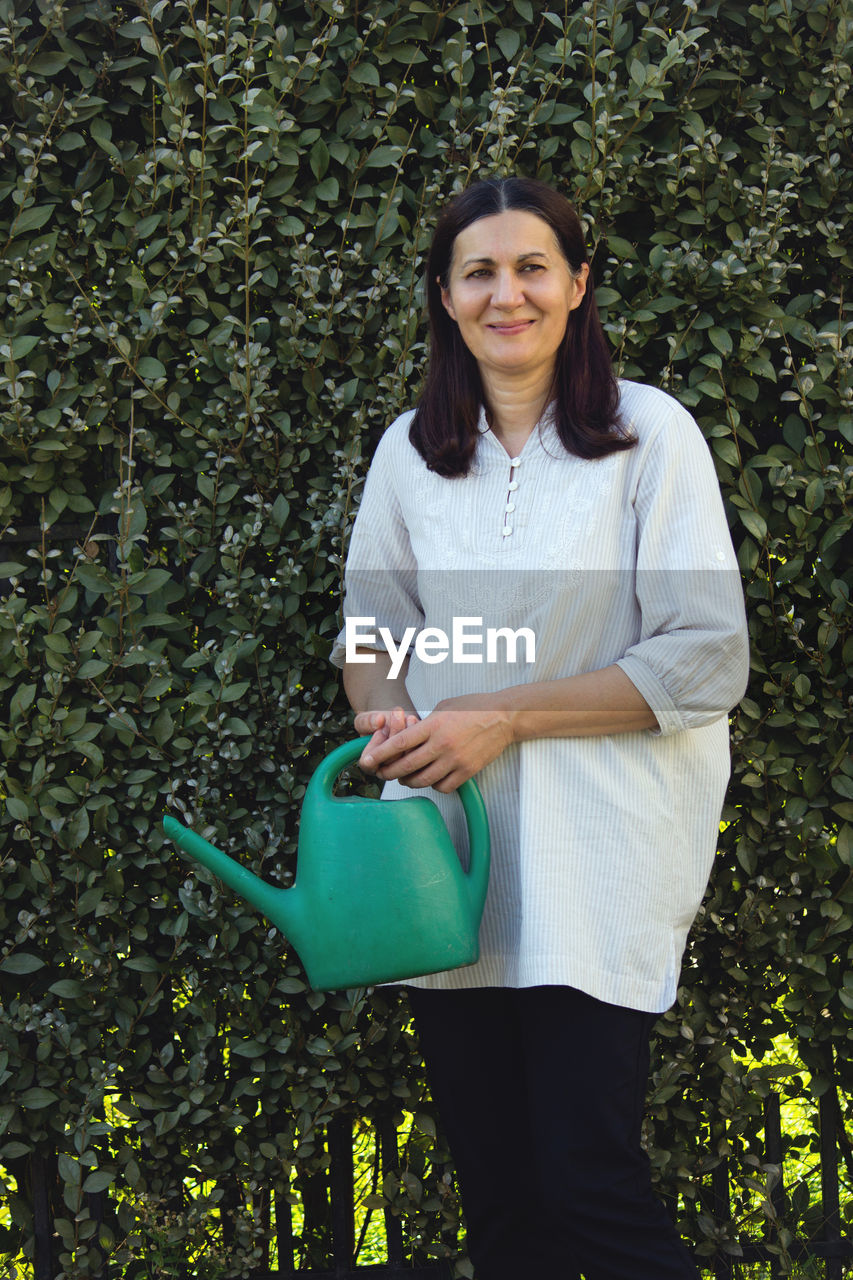 Smiling woman holding watering can while standing against plants
