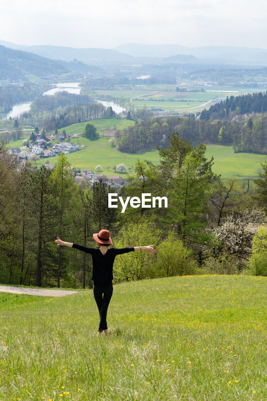 Young skinny woman with open arms and hat stands on a hill in nature of switzerland in springtime.