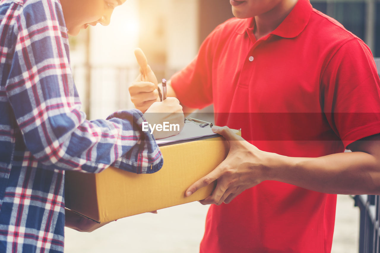 Midsection of female customer signing receipt while receiving package from delivery man outdoors