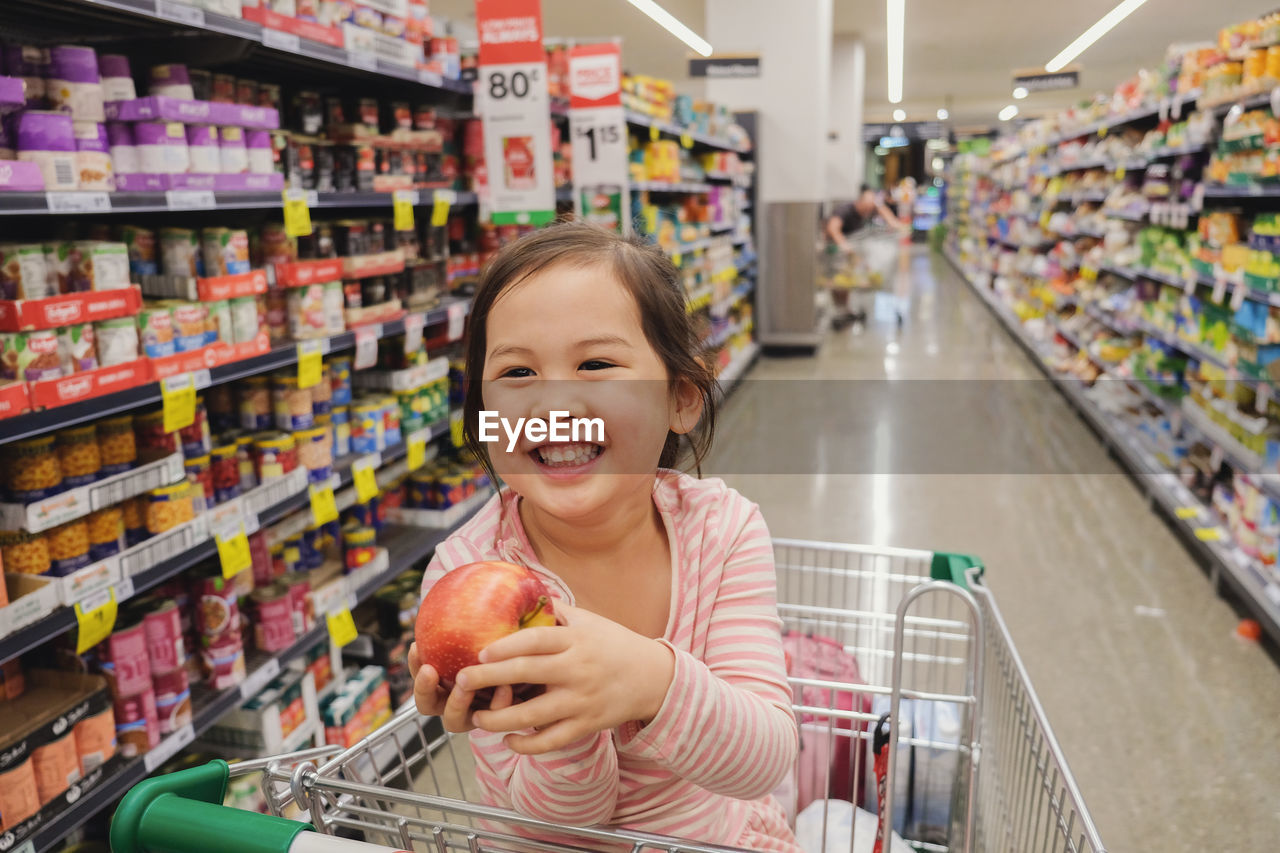 Happy girl holding apple while sitting in shopping cart at store